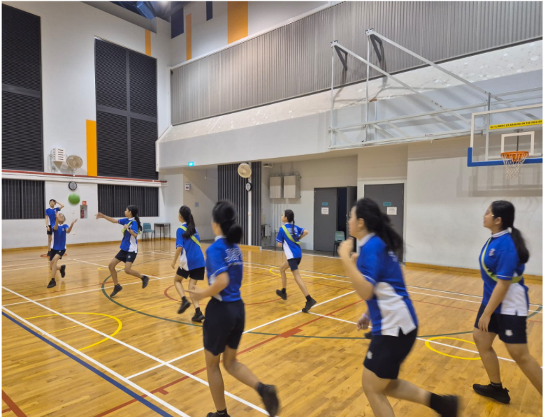 Students in blue uniforms play basketball in an indoor gymnasium with a wooden court and visible hoop.