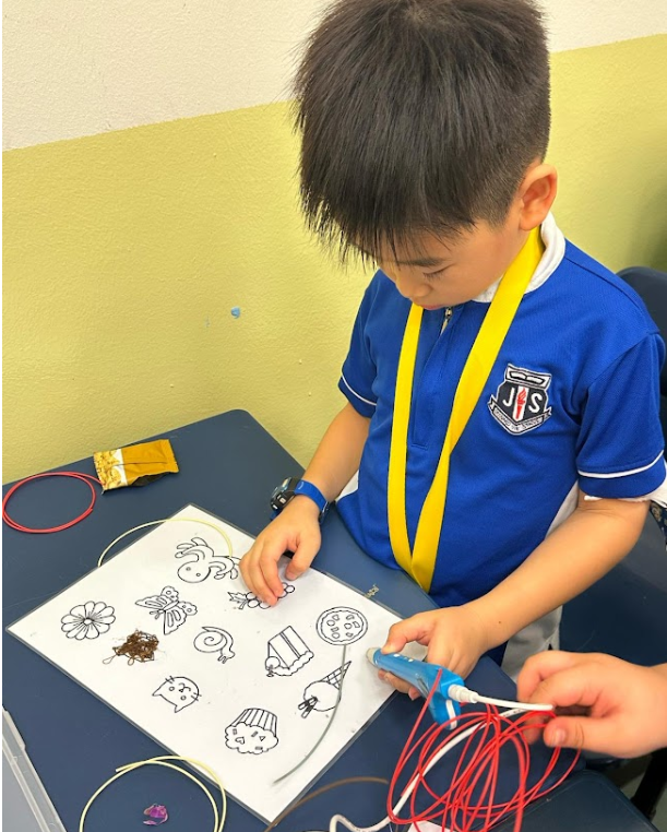 Young boy in blue school uniform using a glue gun on a sheet of cartoon drawings at a desk.