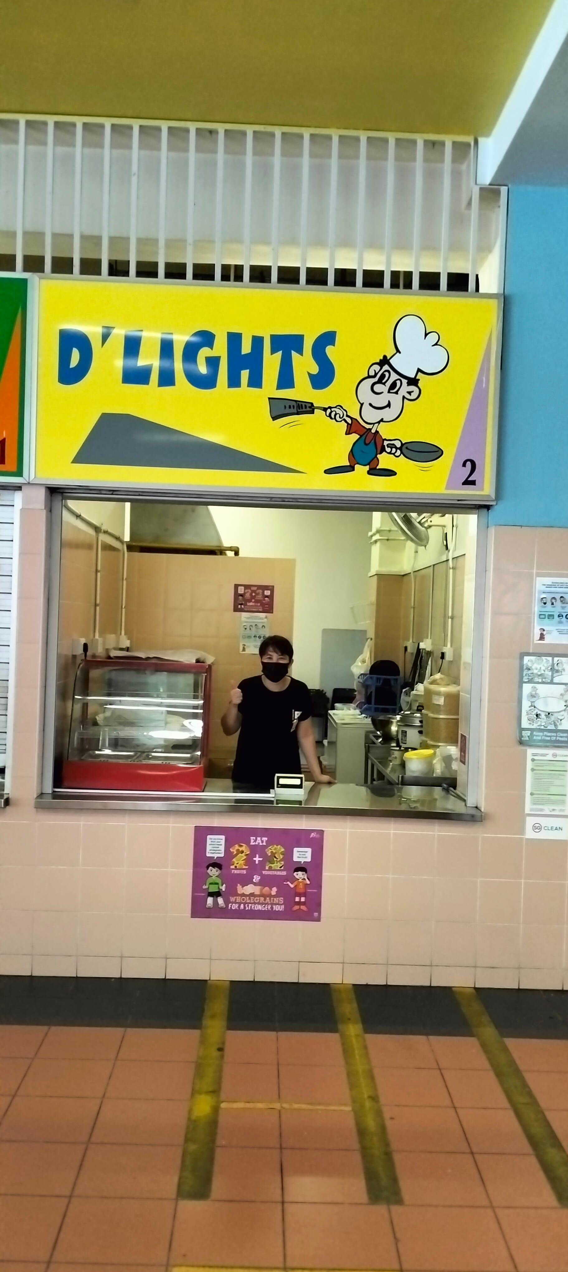 A food stall named D'Lights with a yellow sign and a masked worker in black standing behind the counter.