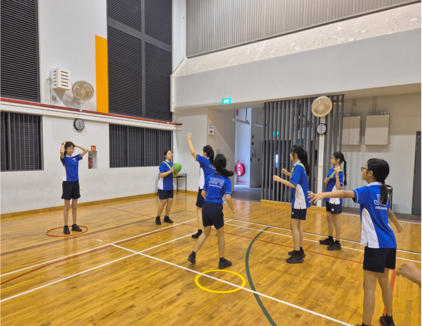 Students in blue uniforms practice ball skills on a wooden indoor court with marked lines.