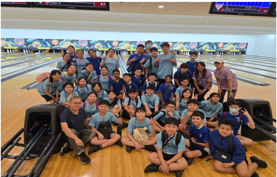 Large group of children and adults in blue shirts posing together at a bowling alley with lanes visible behind them.