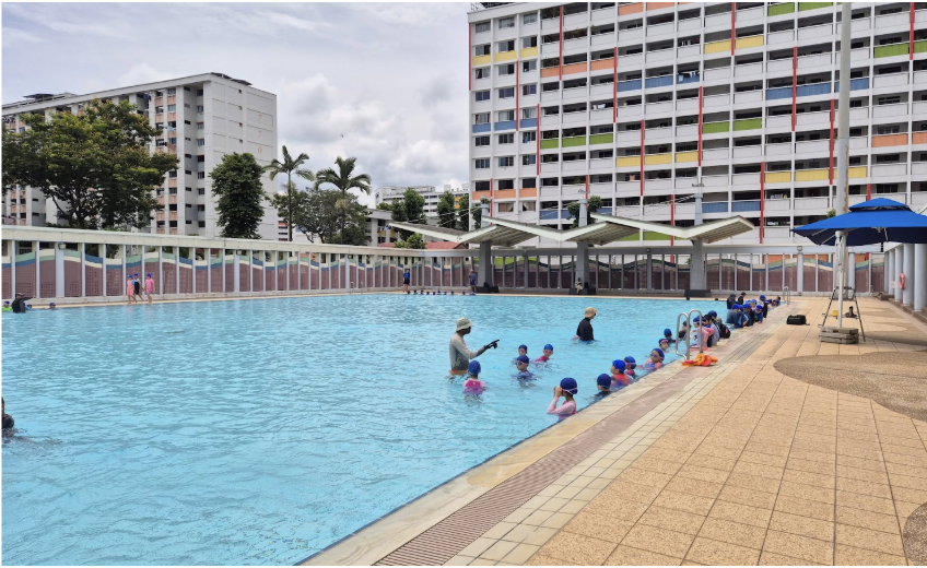 Children in blue swim caps receive swimming lessons in an outdoor pool surrounded by colorful apartment buildings.