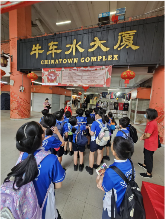 Students in blue uniforms gather at the entrance of Singapore's Chinatown Complex under a large sign.