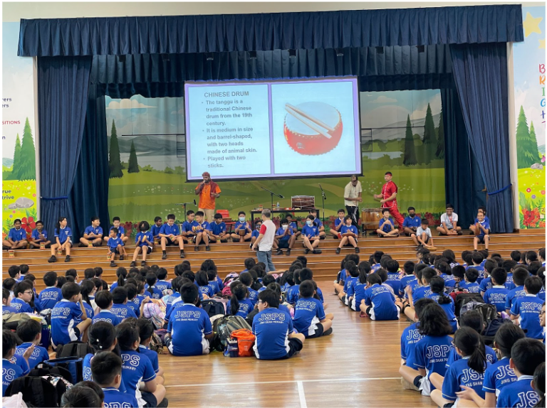 Students in blue JSPS uniforms sit on a gym floor watching a school assembly with a Chinese drum slideshow on stage.