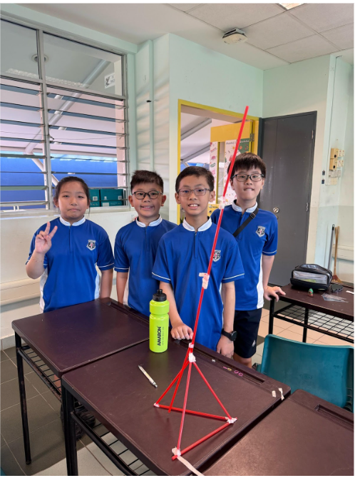 Four students in blue uniforms stand behind a desk displaying a tall structure built from red straws.