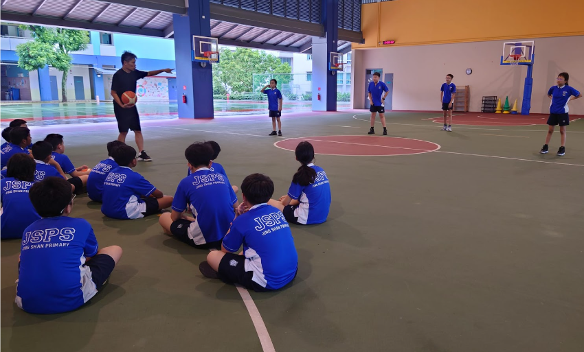 A coach holding a basketball instructs JSPS-uniformed students sitting on an indoor court.