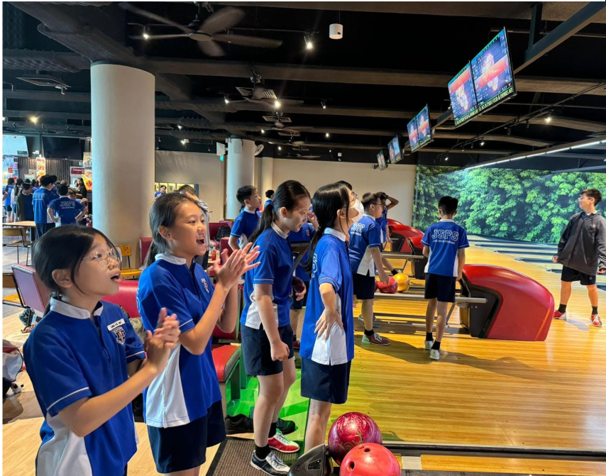 Children in blue school uniforms cheer and clap at a bowling alley with lanes, balls, and overhead screens visible.