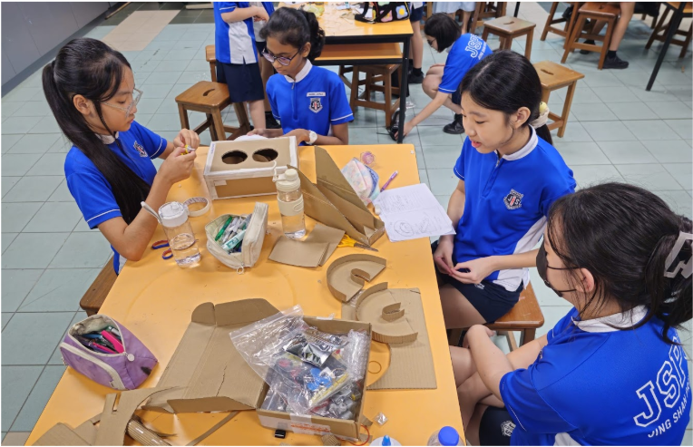 Students in blue uniforms work together at a table covered with cardboard pieces and craft supplies.