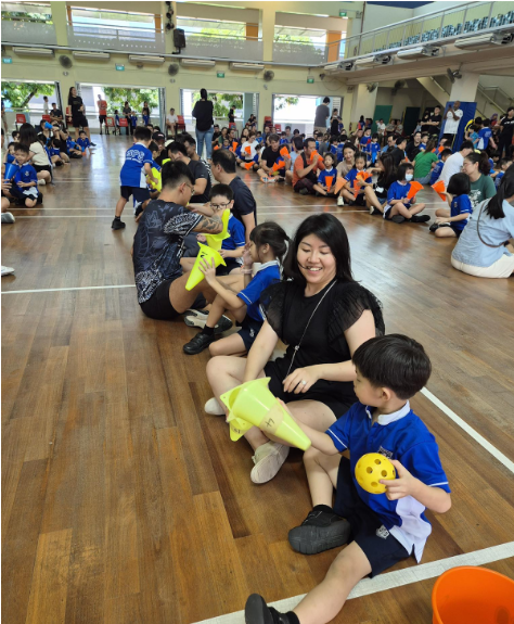 A smiling woman in black sits on a gymnasium floor among many children in blue uniforms during a school event.