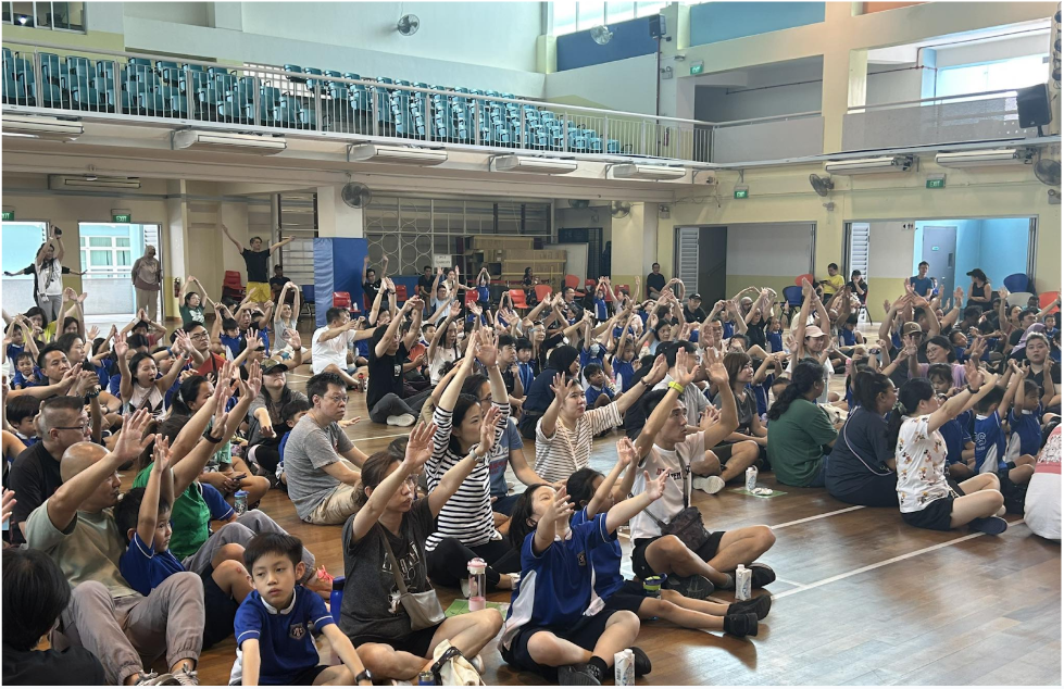 Large crowd of children and adults sitting on a gym floor with hands raised during a school event.
