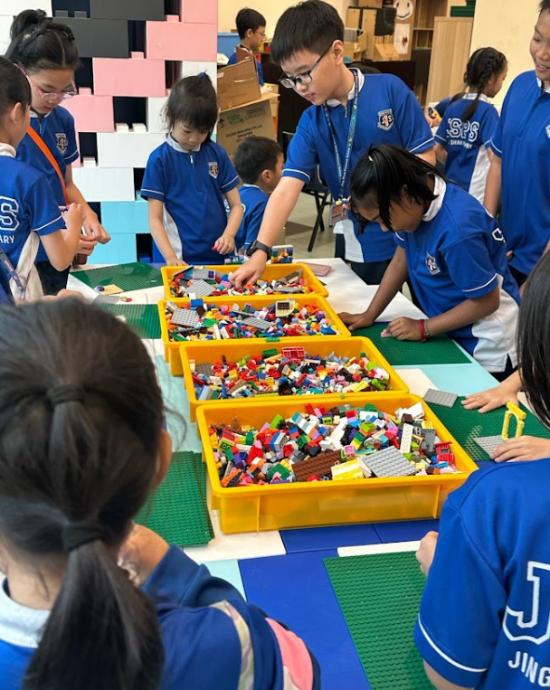 Children in blue school uniforms gather around a table with yellow trays filled with colorful LEGO bricks.