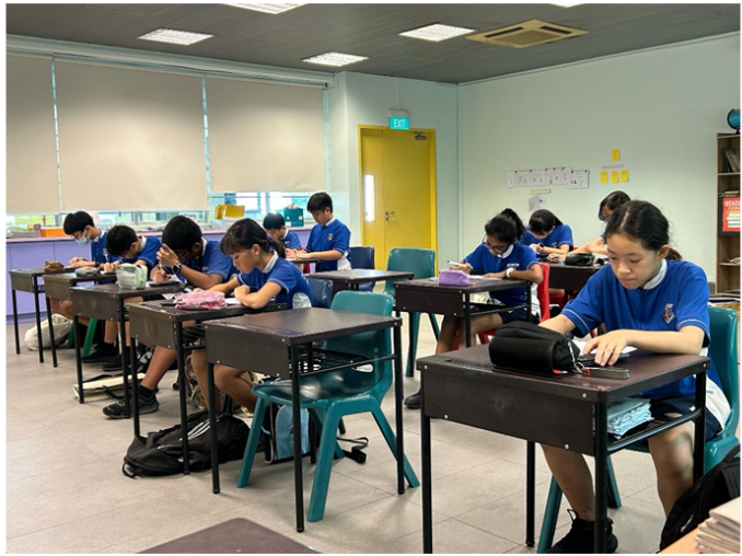 Students in blue uniforms sit at individual desks in a classroom, working quietly on tasks.