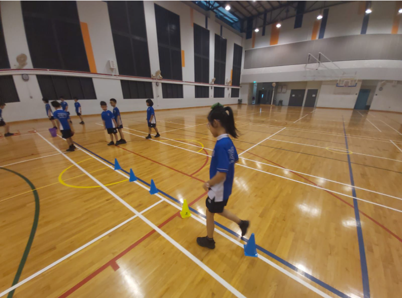 Children in blue uniforms doing a cone drill on a wooden indoor sports court with a net in the background.