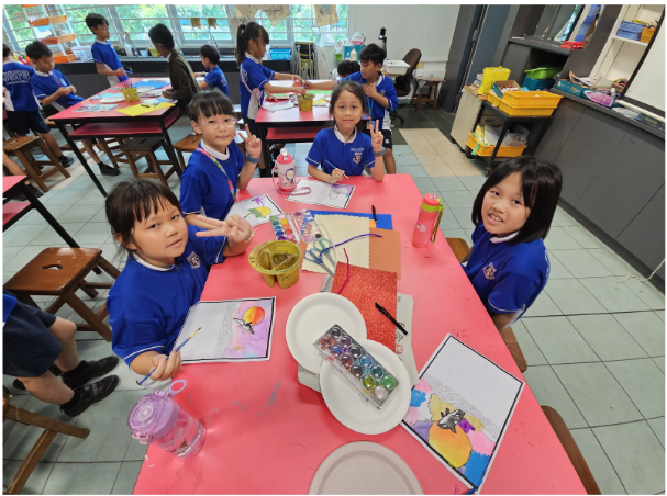 Children in blue uniforms sit at a pink table doing art with paints and colorful drawings in a classroom.