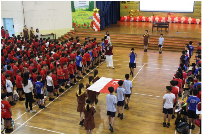 Children in red and blue uniforms stand in rows in a school hall as students carry a Singapore flag.