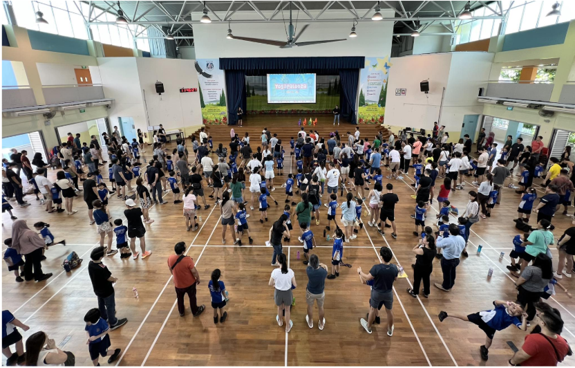 Large indoor school hall filled with crowds of adults and children facing a stage with a projection screen.