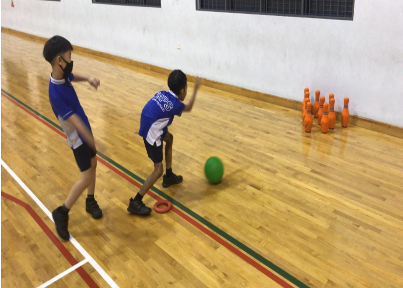 Two boys in blue jerseys bowl a green ball toward orange pins on an indoor wooden court.