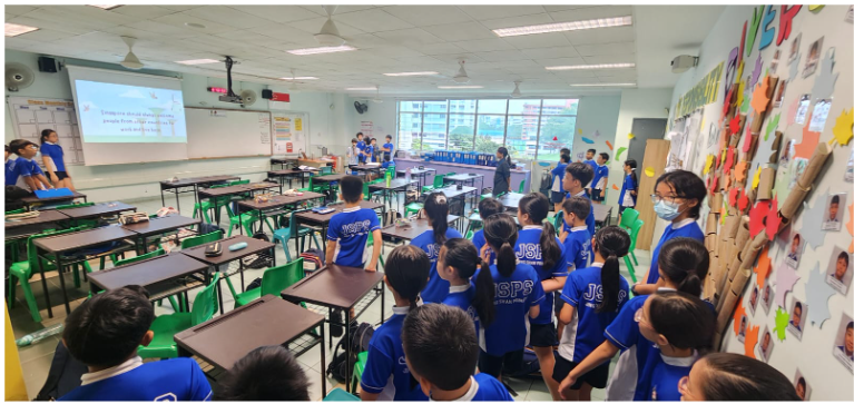 Students in blue JSPS uniforms stand in a classroom facing a projected screen at the front.