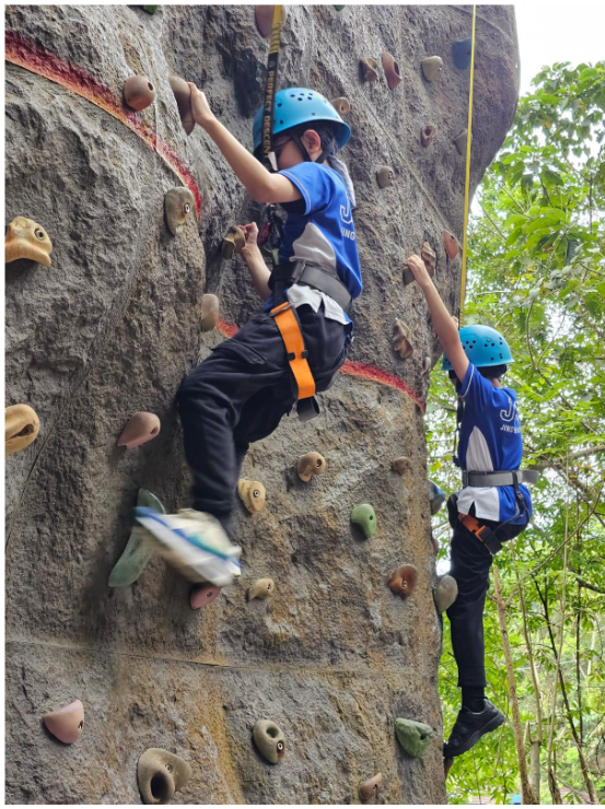 Two children wearing blue helmets and harnesses climb an outdoor rock wall with holds.