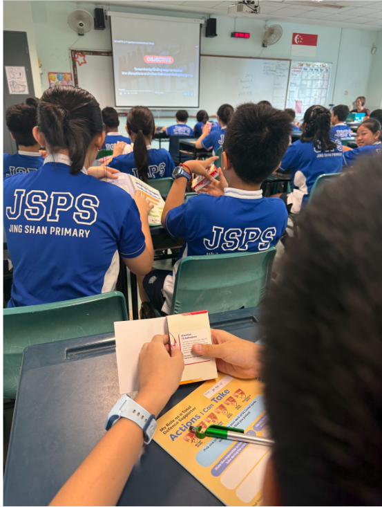 Students in blue Jing Shan Primary School uniforms sit in a classroom facing a projector screen.