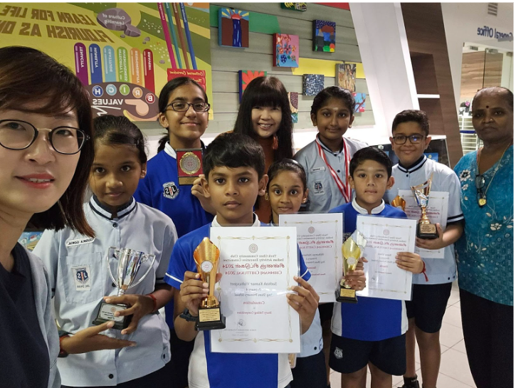 Group of school children and adults posing with trophies and certificates inside a colorful school hallway.