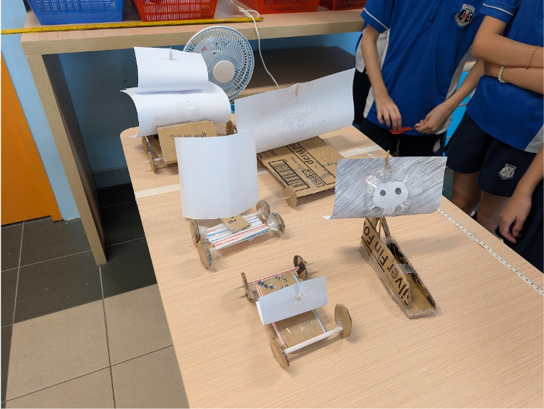 Students in blue uniforms stand beside a table holding cardboard and paper sail-powered toy vehicles near a small fan.