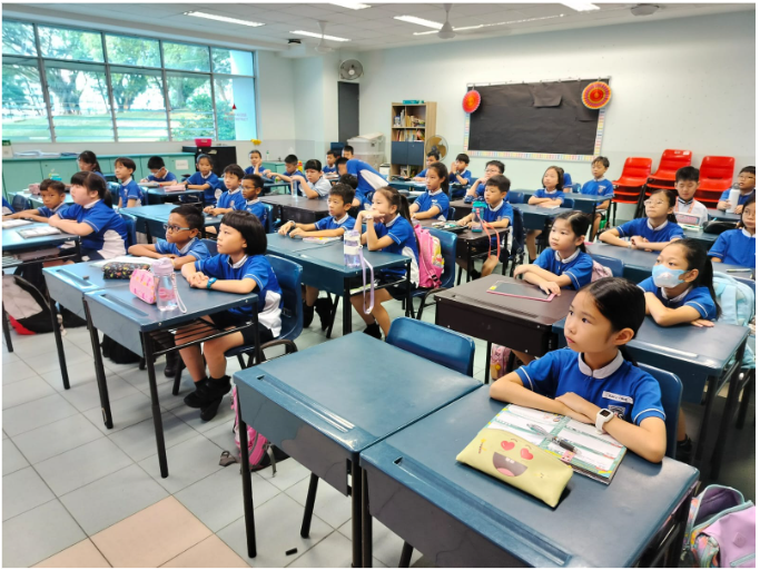 Young students in blue uniforms sit at desks in a bright classroom, facing the front attentively.
