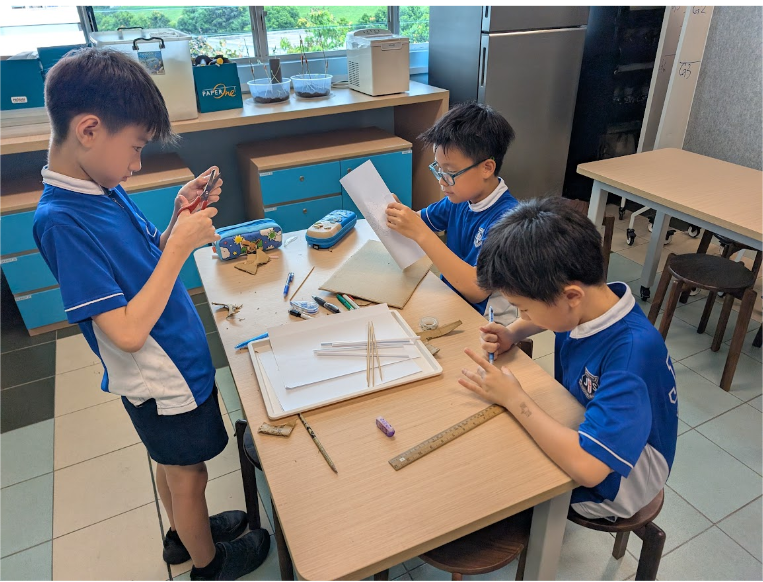 Three boys in blue uniforms work on a craft project at a table with paper, rulers, and pencils.