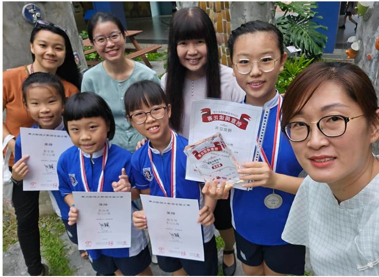 Children in blue uniforms hold certificates and medals while smiling with adult women outdoors.