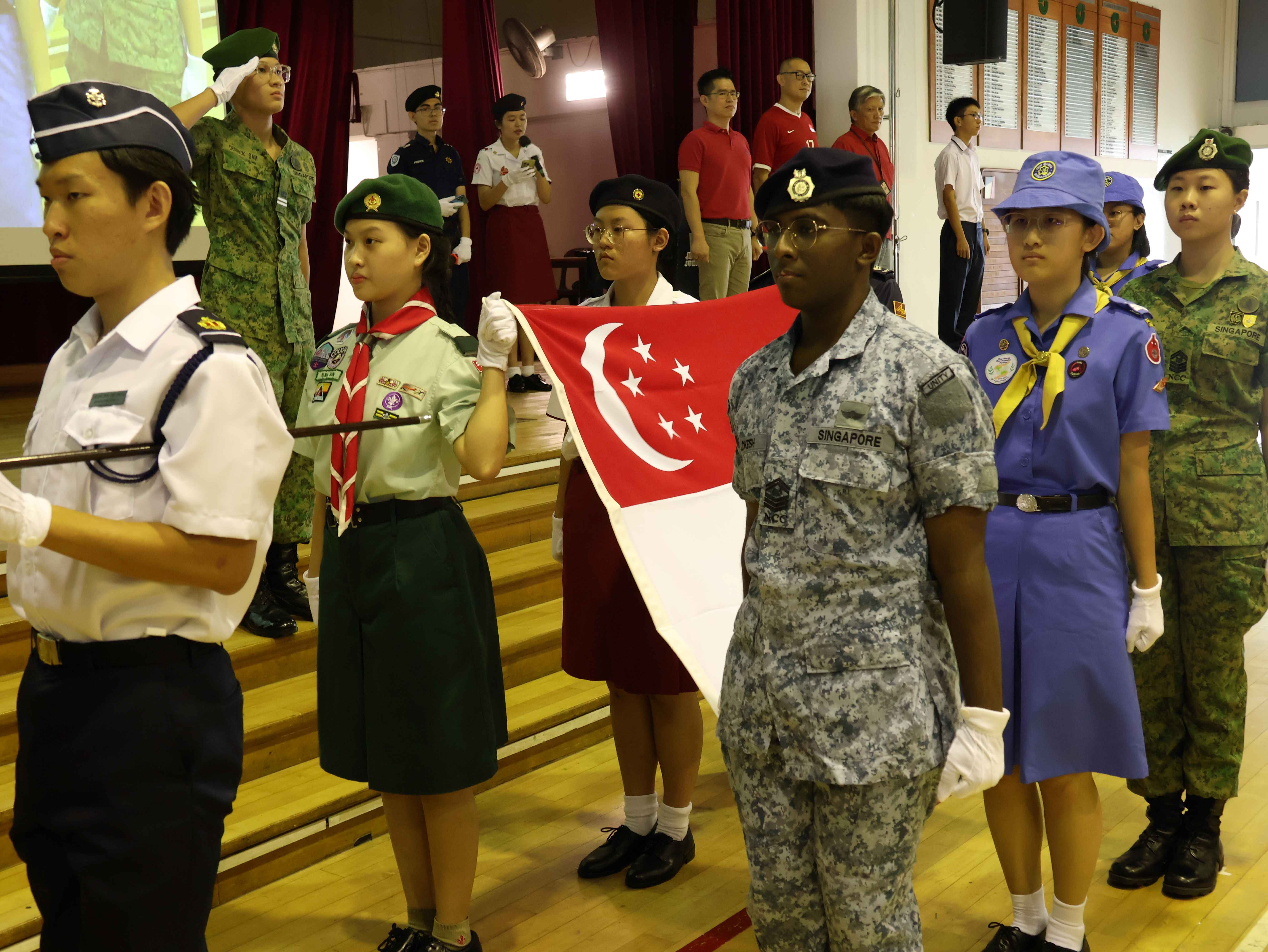 Students in various Singapore uniformed group attire stand at attention during a flag ceremony indoors.