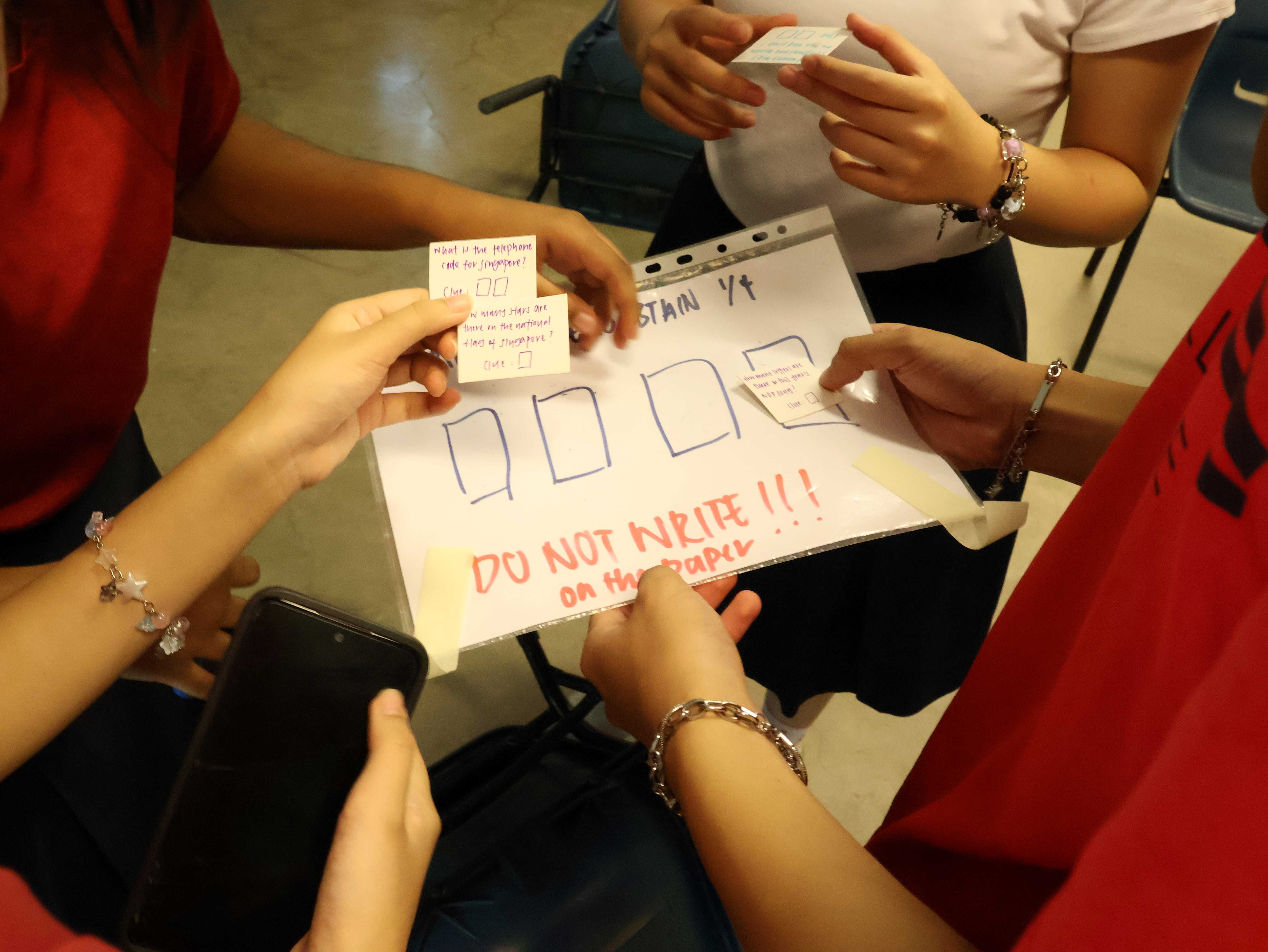 Several hands holding a sheet with drawn boxes and red text reading "DO NOT WRITE on the paper" with small clue cards.