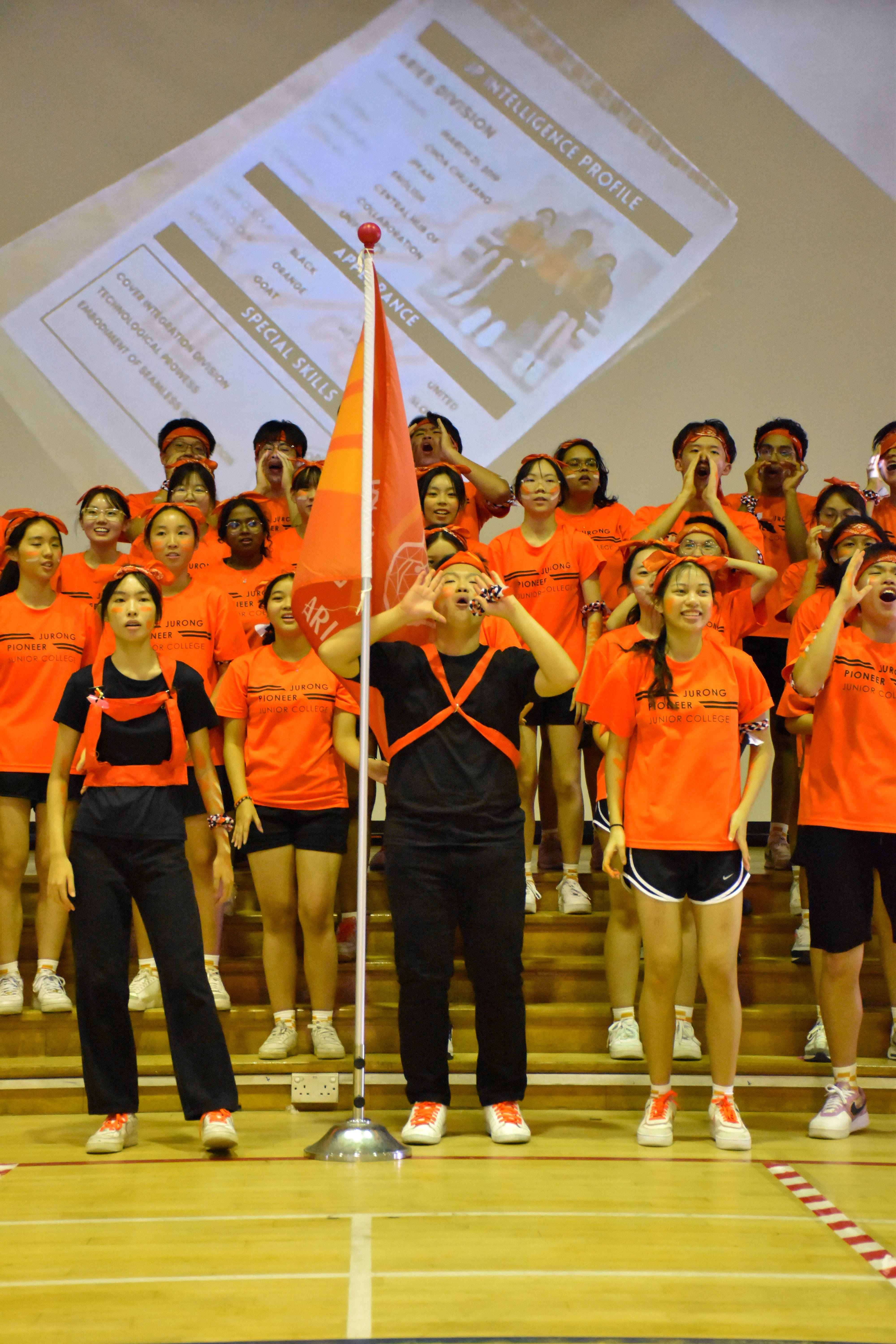 A large group of students in orange Jurong Pioneer Junior College shirts cheering on tiered steps with an orange flag.