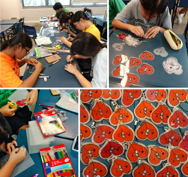 Students craft red heart-shaped keychains at blue tables using colored pencils and scissors in a classroom.