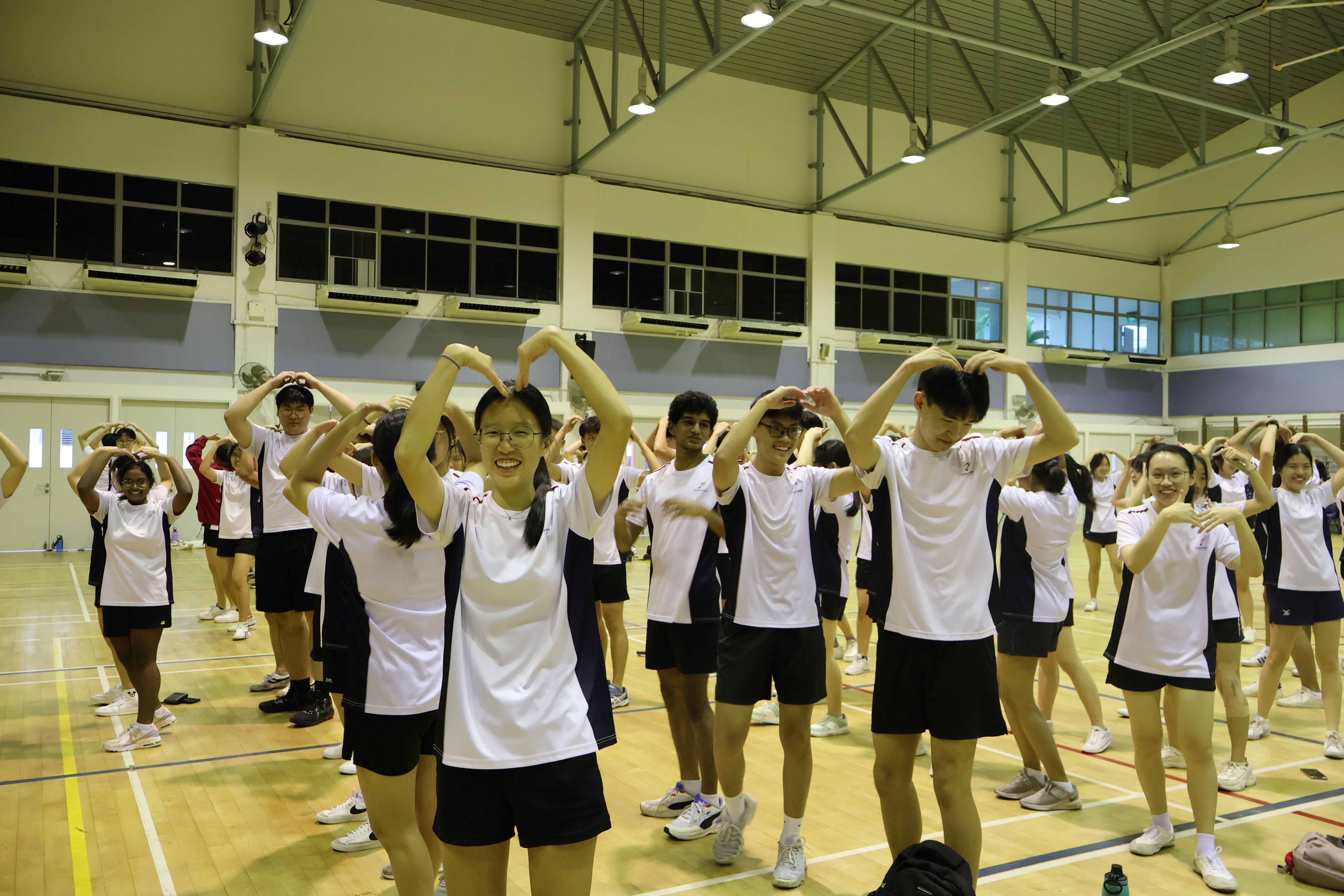 Students in white and navy uniforms form heart shapes with their arms overhead inside a school gymnasium.
