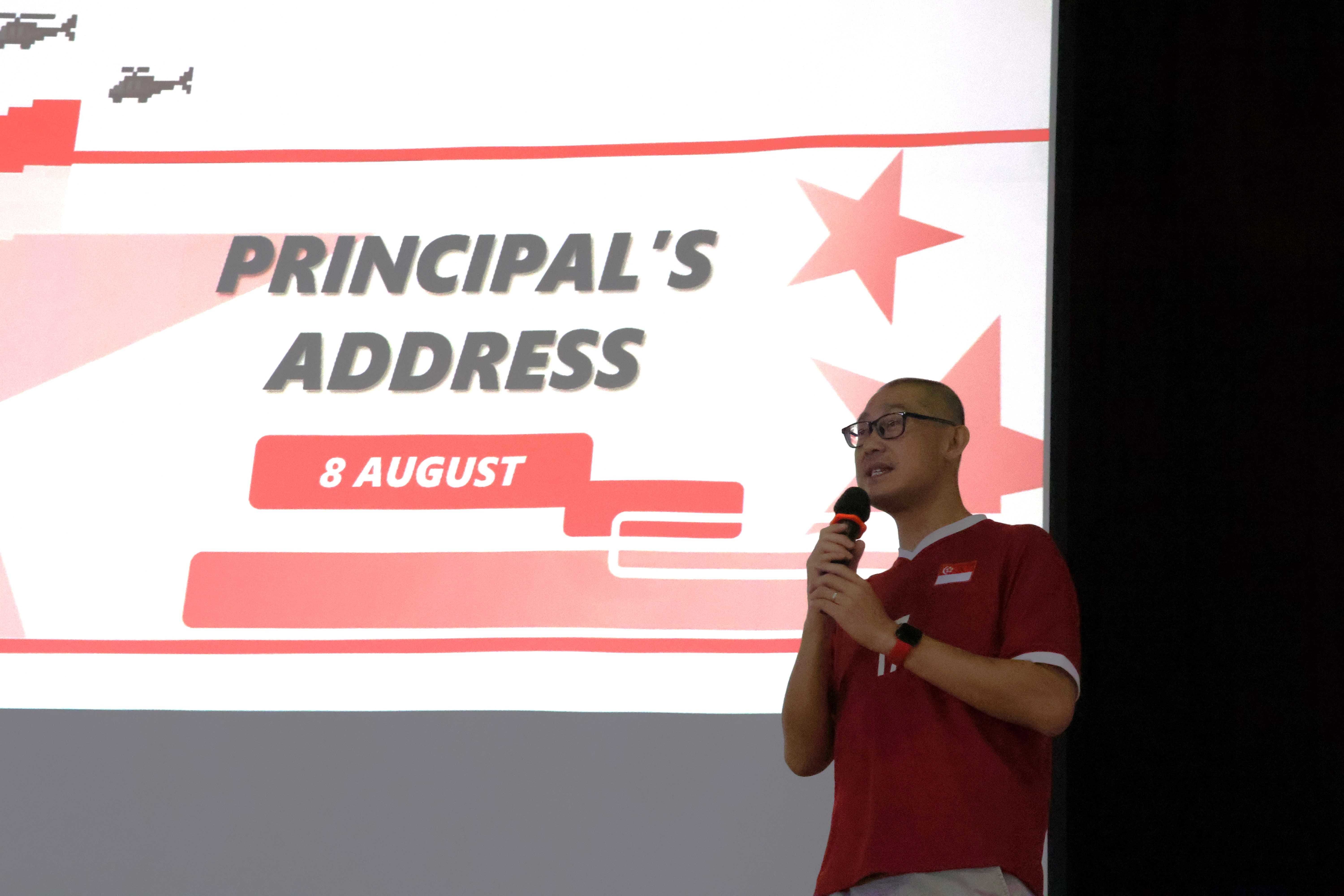 Man in red shirt holding a microphone speaks in front of a slide reading Principal's Address, 8 August.