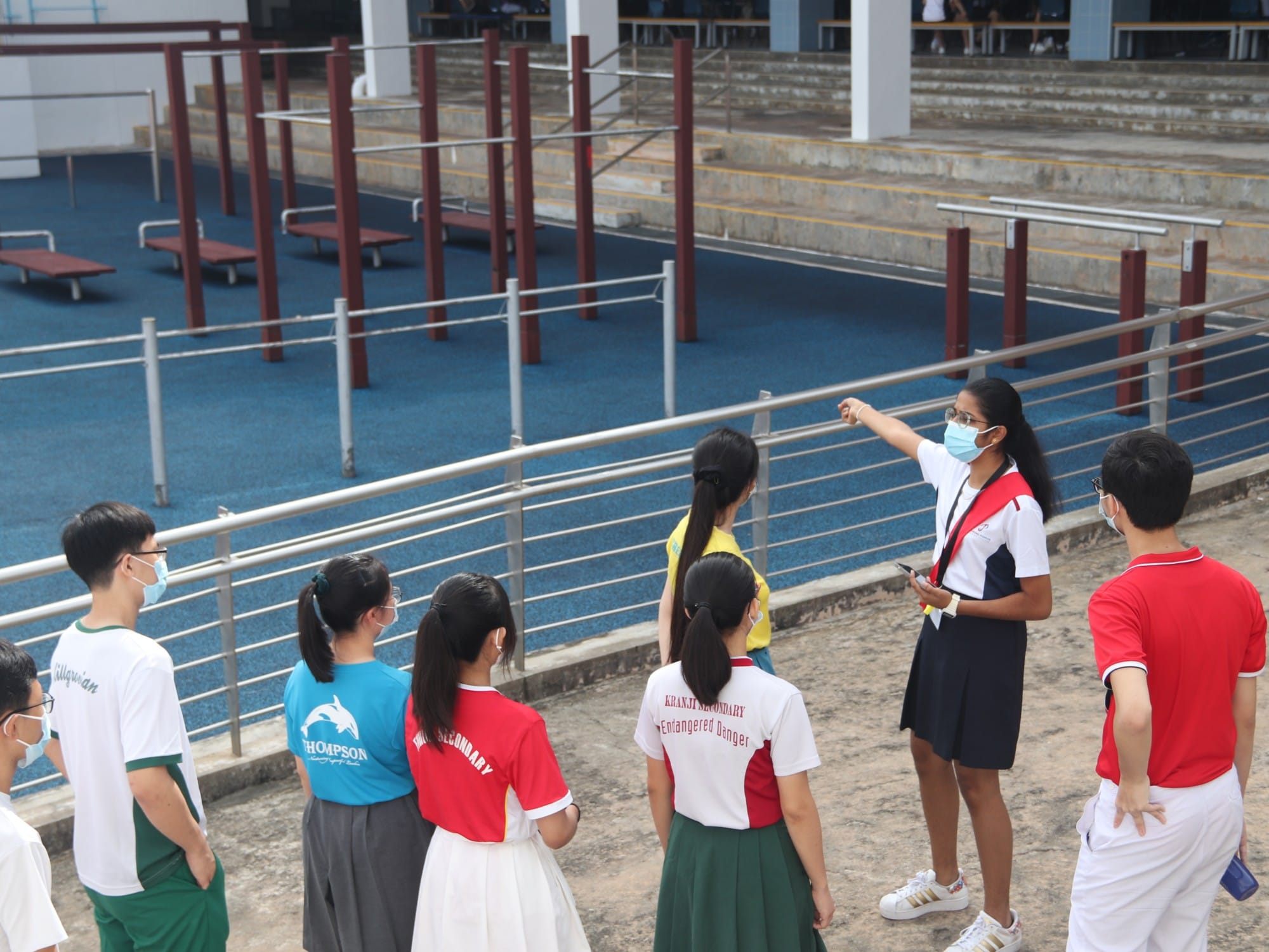A masked student guide points toward a blue courtyard while leading a group of masked students outdoors at a school.