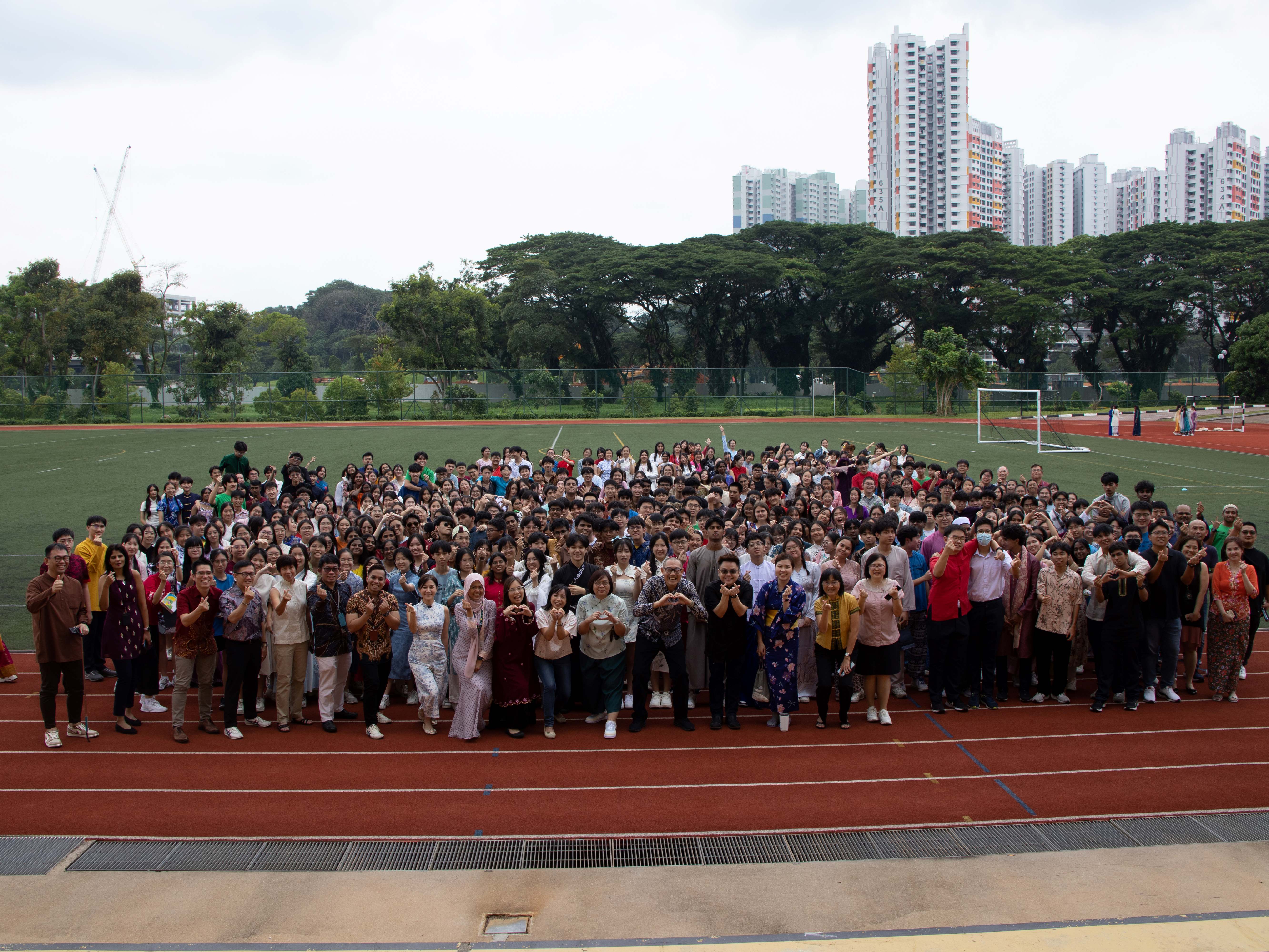 Large group of students in colorful traditional attire posing on a school running track with a soccer field behind them.