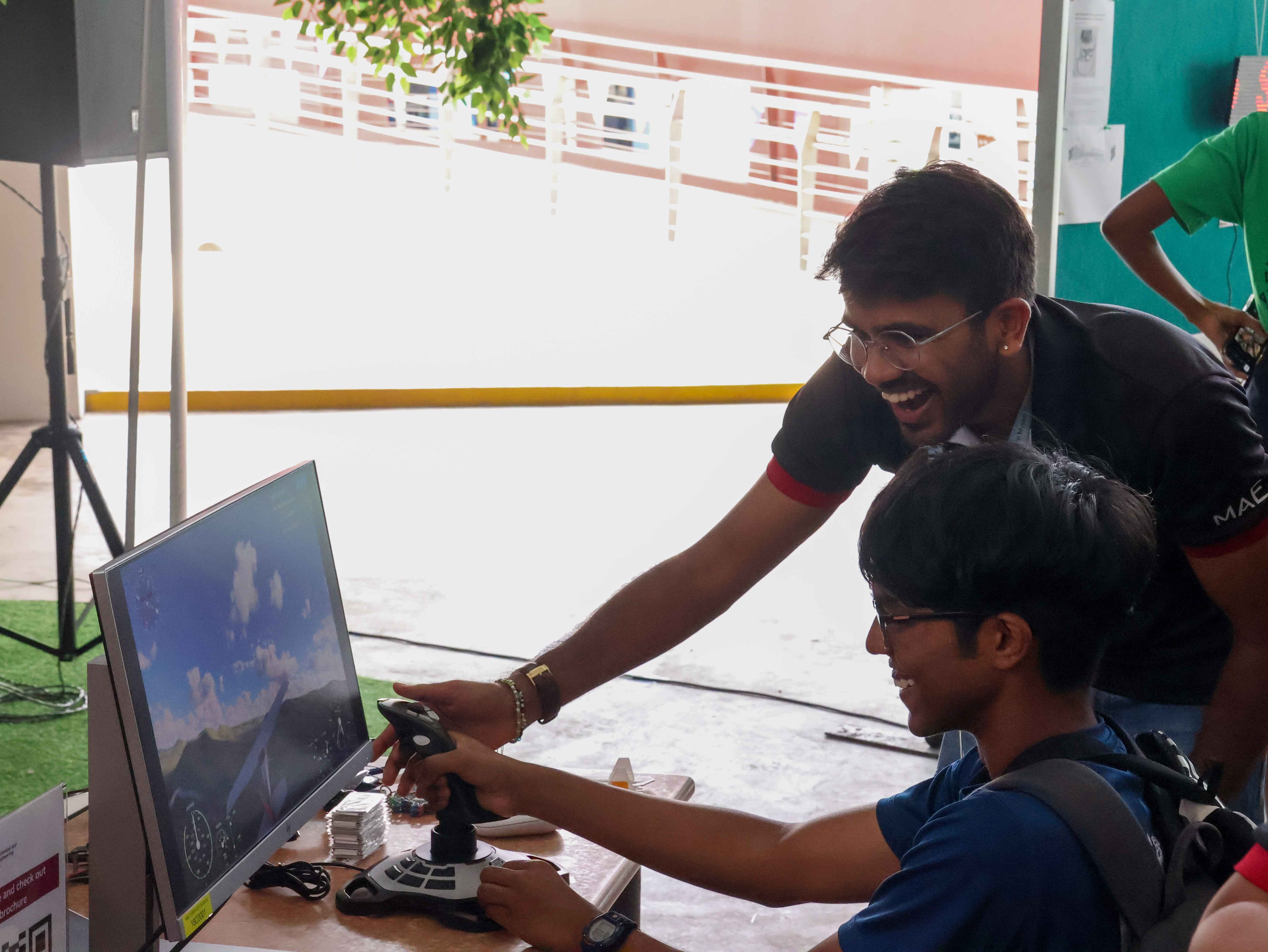 Two young men smile while using a joystick at a flight simulator display on a monitor at an outdoor event.