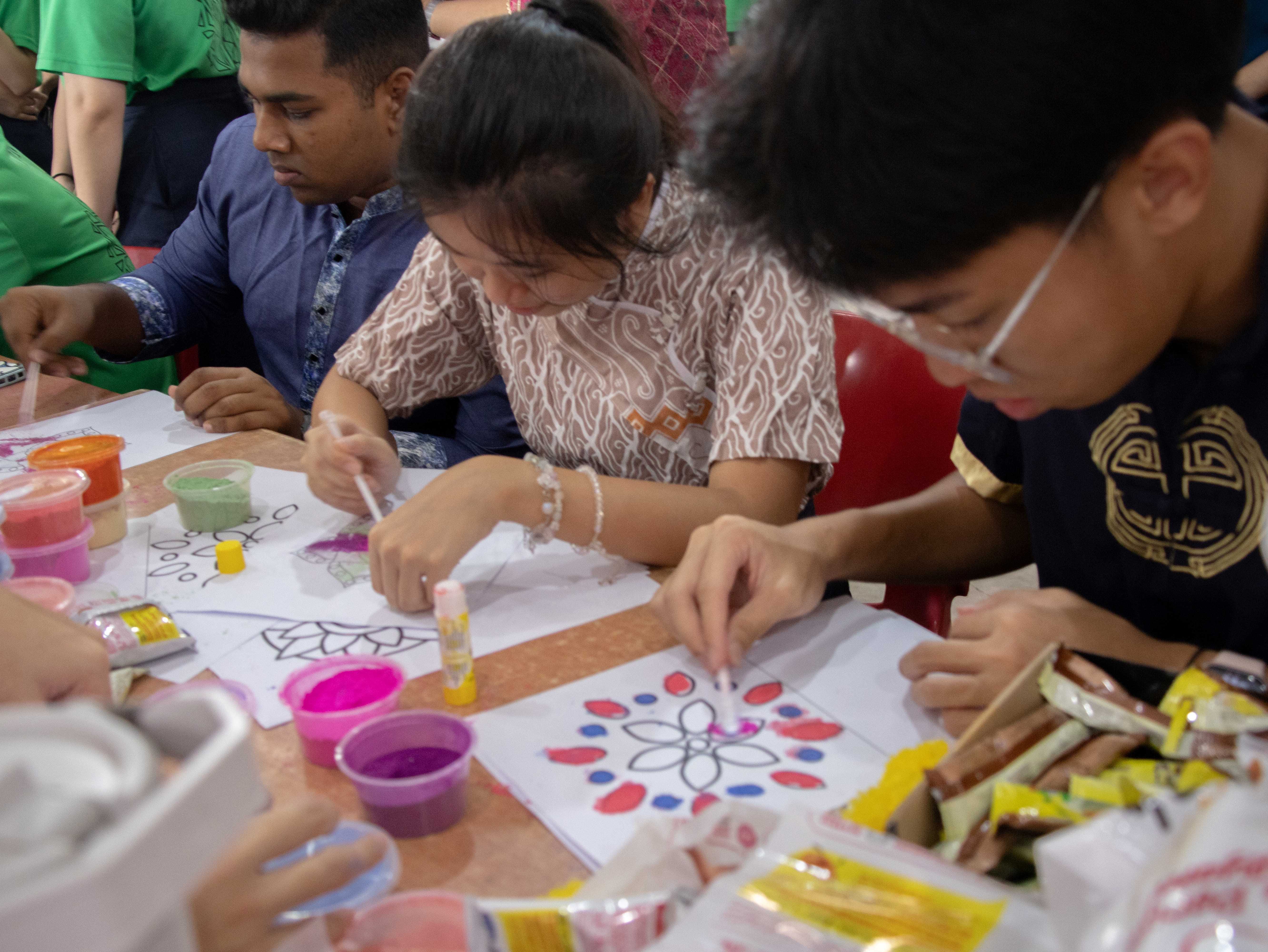 Several people sit at a table painting colorful floral designs on paper using small brushes and cups of paint.