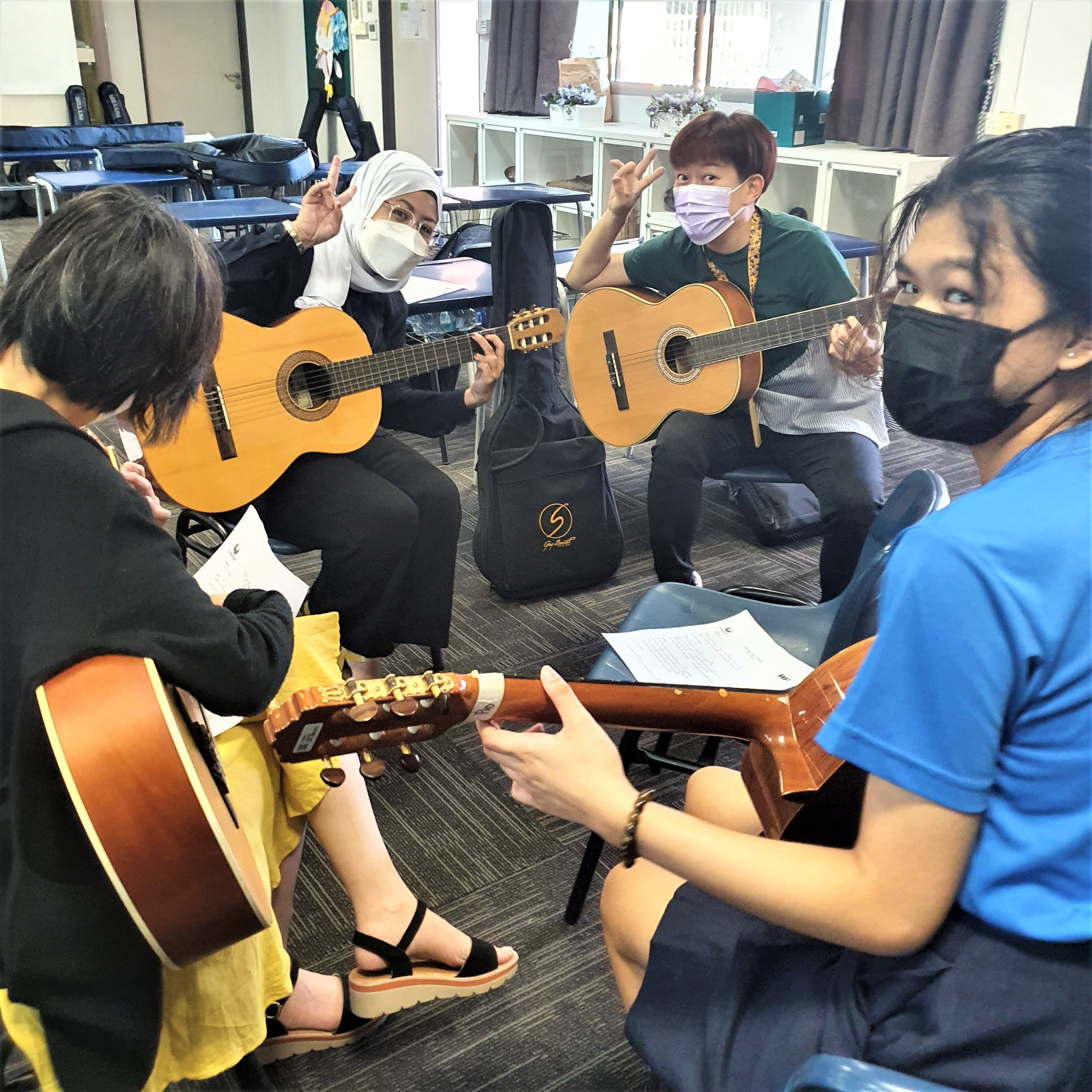 Four masked people sit in a circle holding acoustic guitars in a classroom, two making peace signs.