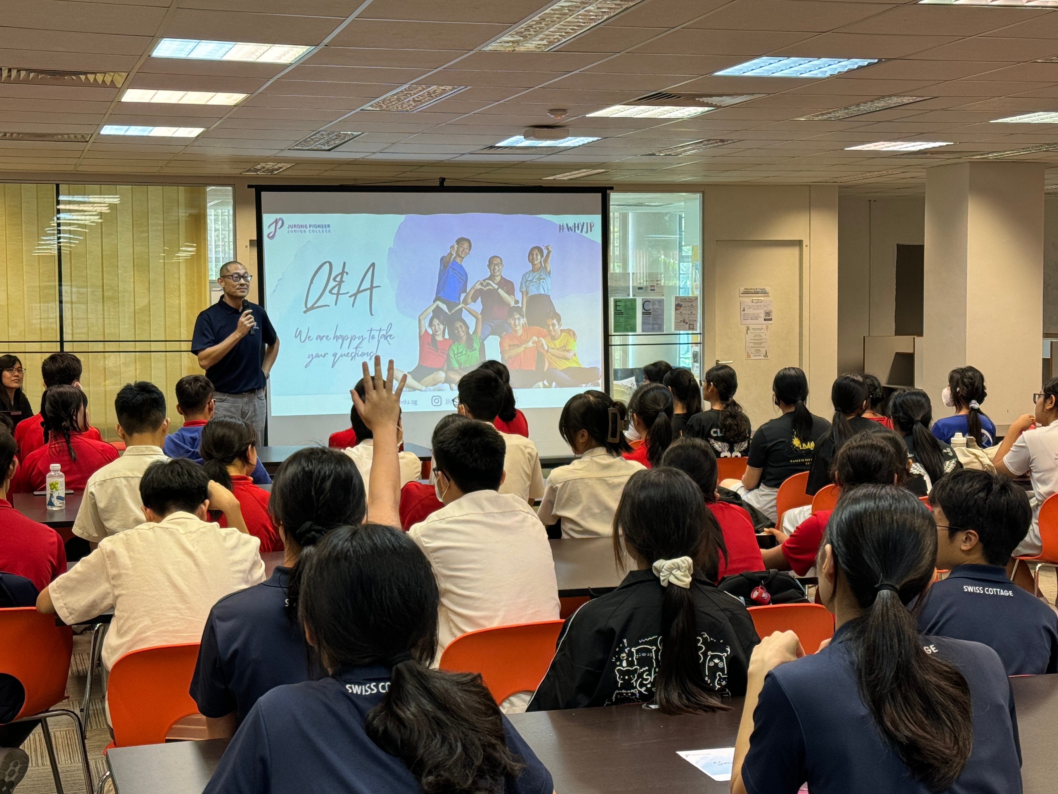A speaker holds a microphone before students in a room, with a Q&A slide from Jurong Pioneer Junior College projected behind.