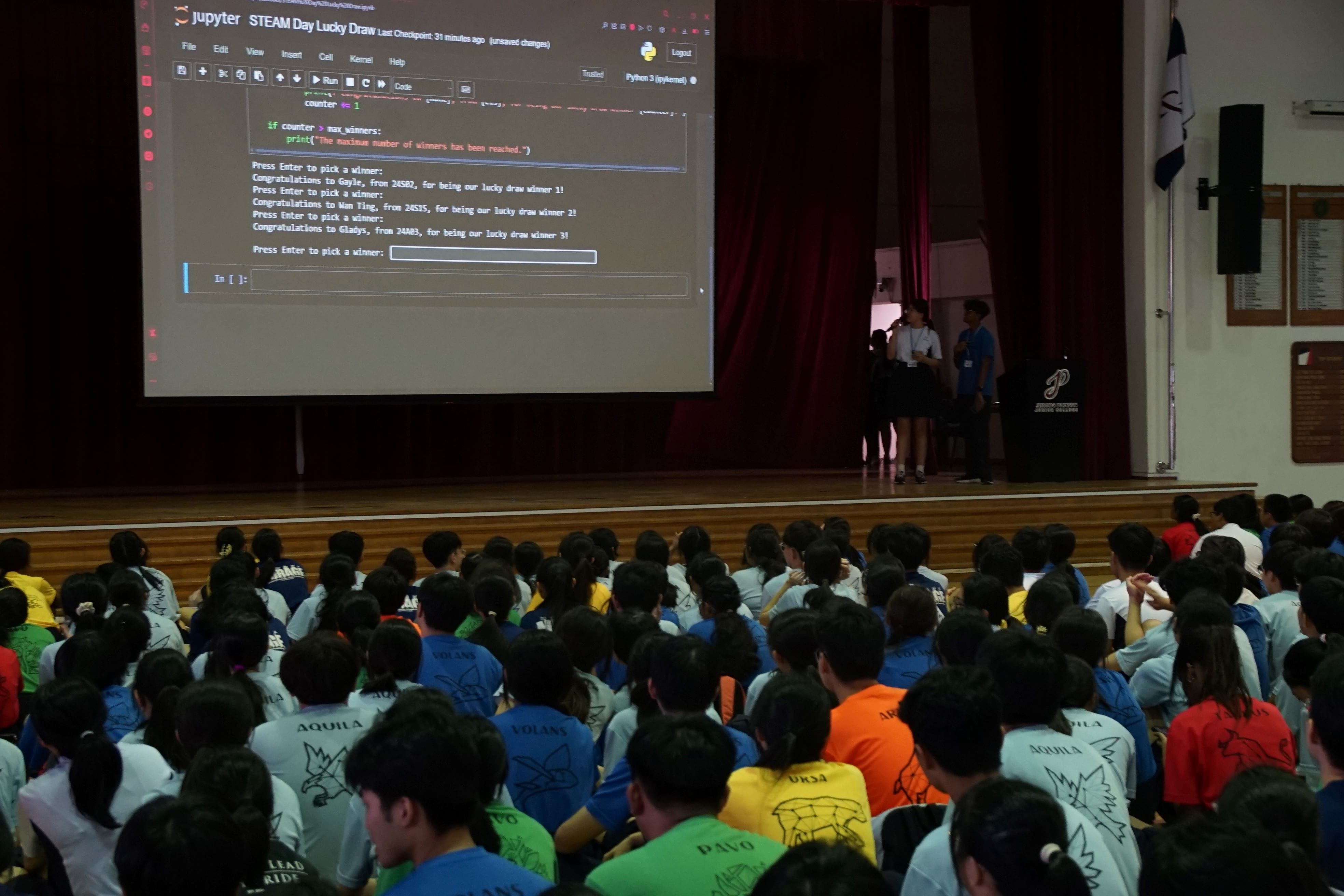 Students in colorful shirts sit in a hall watching a Jupyter notebook lucky draw program projected on a large screen.