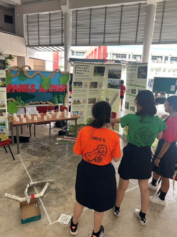 Students in orange and green shirts stand before a nature-themed display board at a school exhibition.