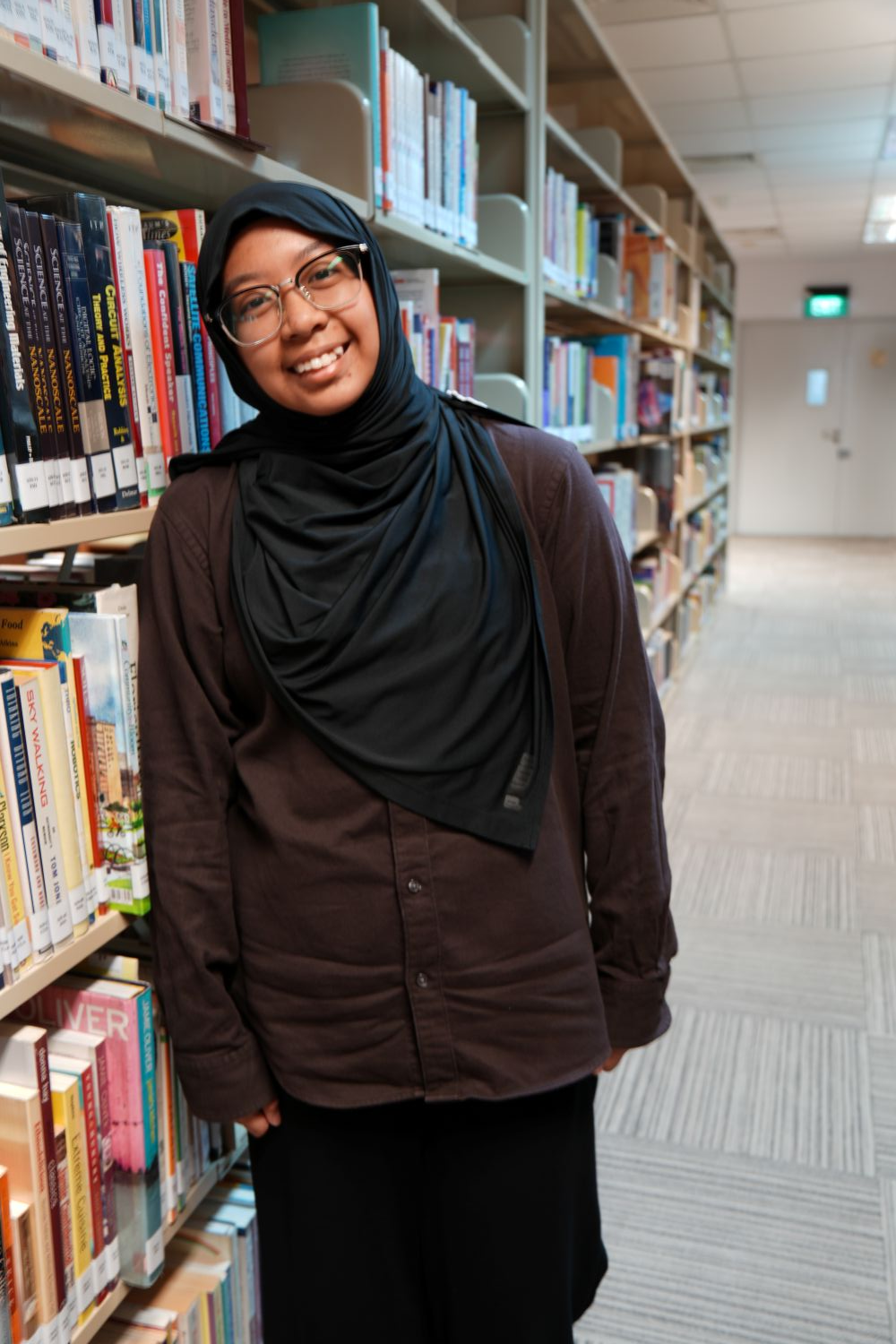 A smiling young woman wearing a black hijab and glasses stands beside library bookshelves.