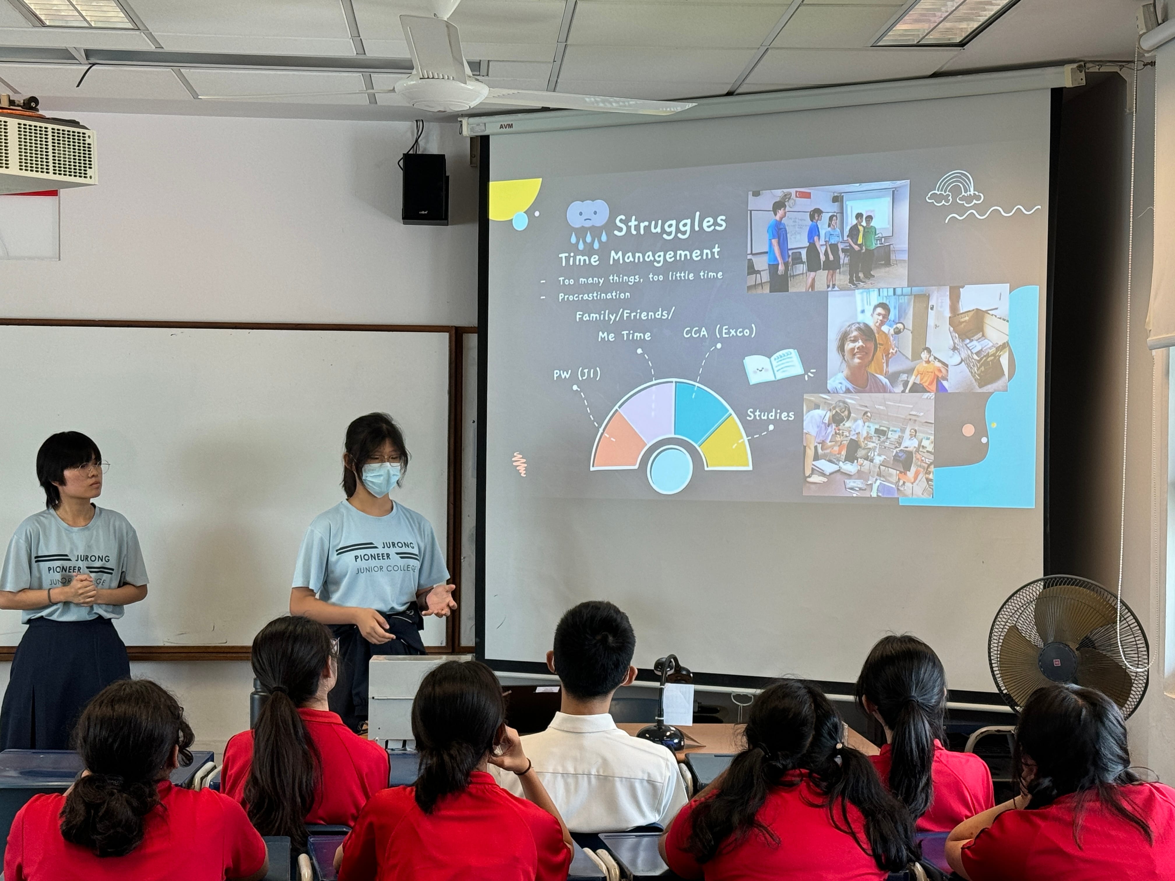 Two students present a slide on Struggles and Time Management to a classroom of students in red uniforms.