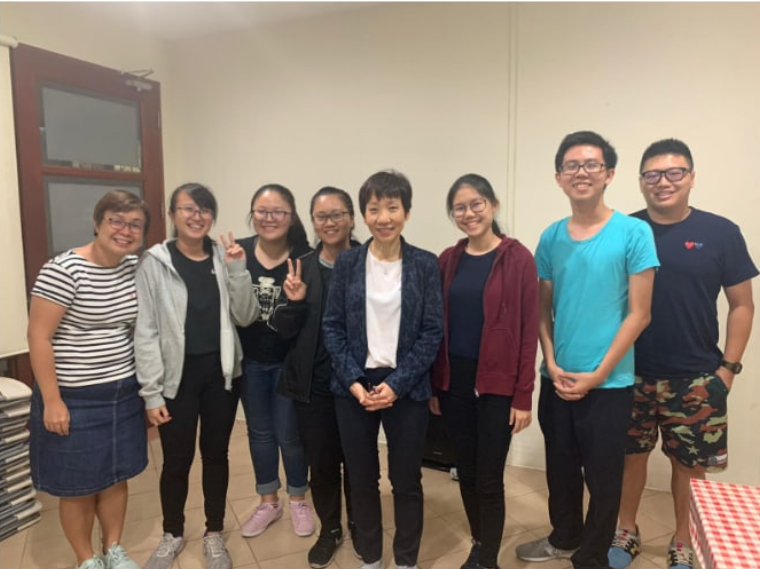 Eight people standing together indoors smiling for a group photo, one person making a peace sign.