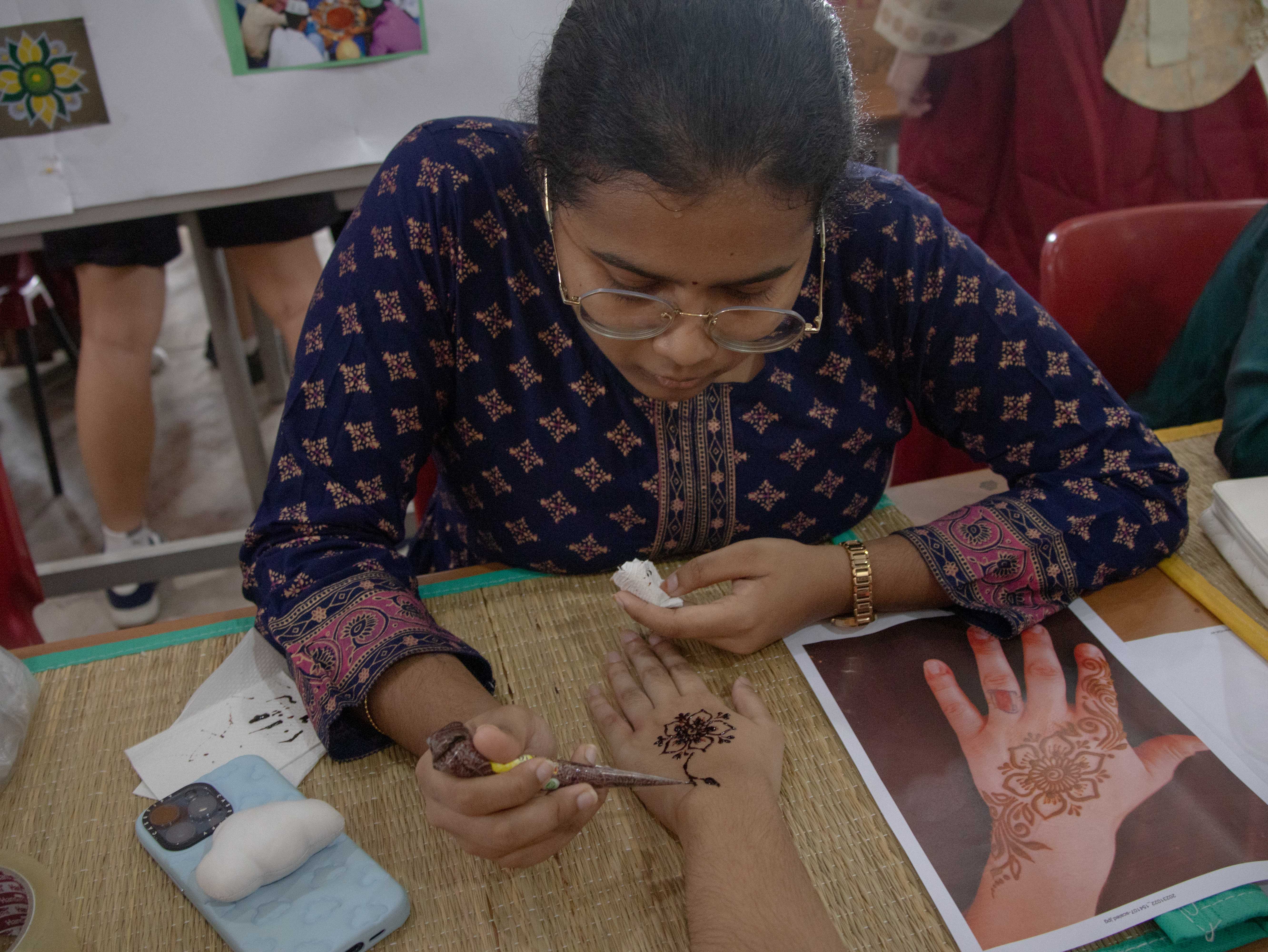 A young woman with glasses applies henna to someone's hand, using a reference photo of a floral design nearby.