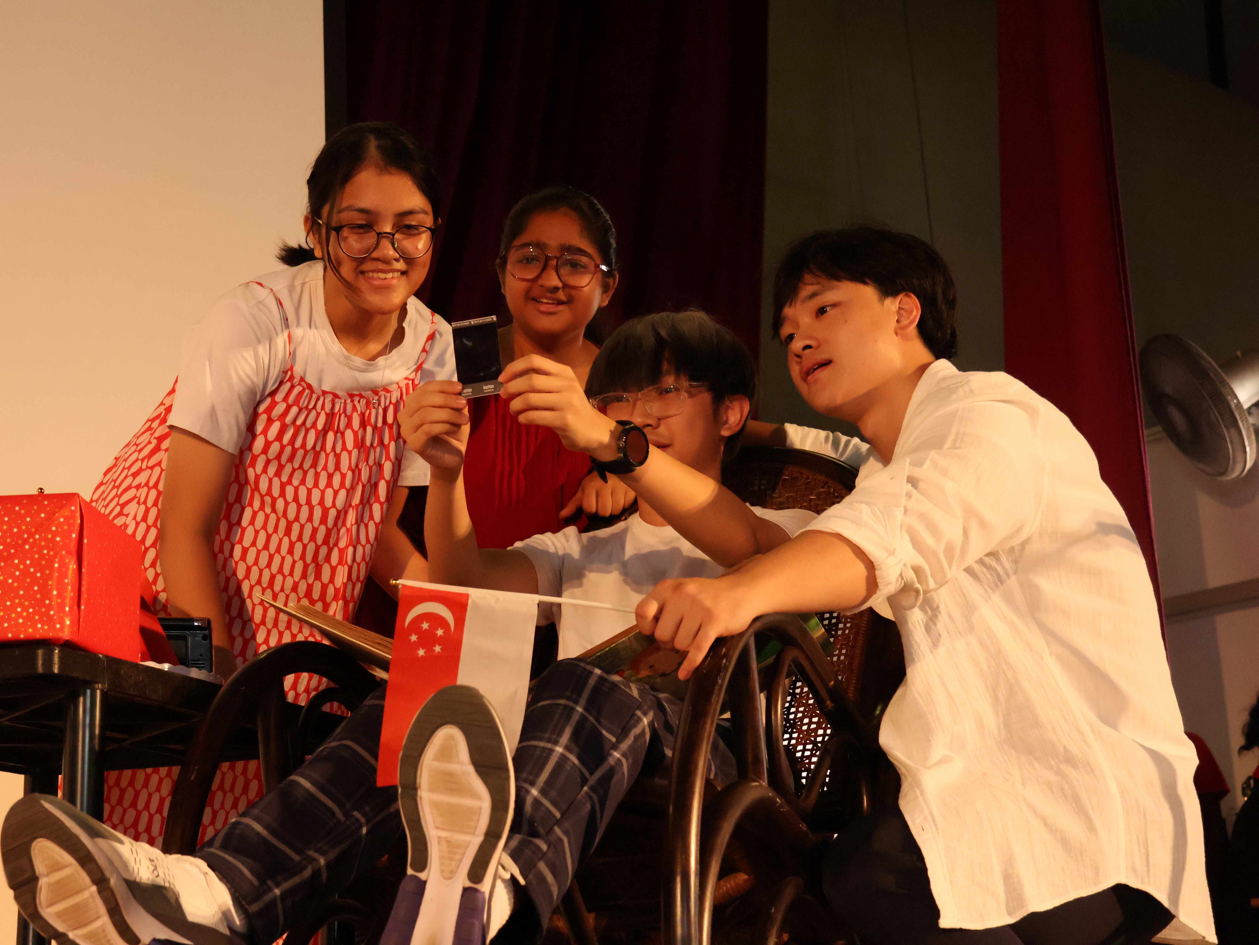 Four teens on a stage, one holding a small Singapore flag and another holding a card, gathered around a wheelchair.