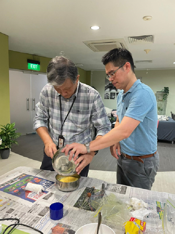 Two men work together over a mixing bowl on a newspaper-covered table indoors.