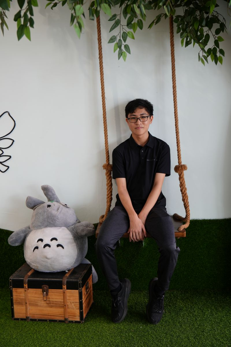 Young man with glasses in black shirt sits on a rope swing beside a gray Totoro plush toy on a wooden chest.